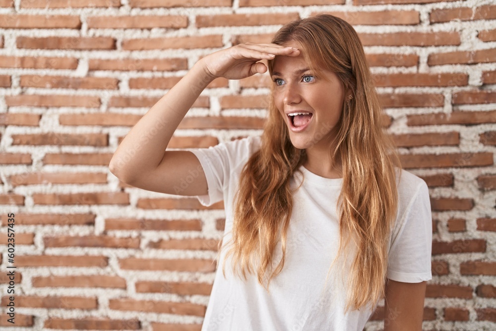 Young caucasian woman standing over bricks wall very happy and smiling looking far away with hand over head. searching concept. Young caucasian woman standing over bricks wall very happy and smiling looking far away with hand over head. searching concept.