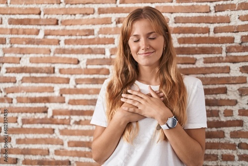 Young caucasian woman standing over bricks wall smiling with hands on chest with closed eyes and grateful gesture on face. health concept.