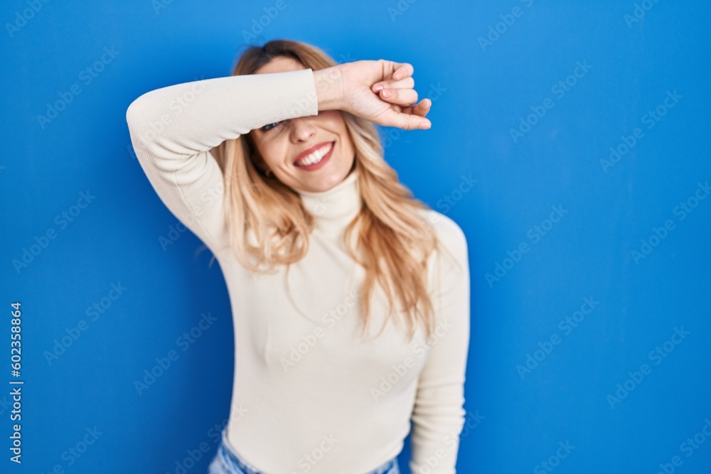 Young caucasian woman standing over blue background covering eyes with arm smiling cheerful and funny. blind concept. Young caucasian woman standing over blue background covering eyes with arm smiling cheerful and funny. blind concept.