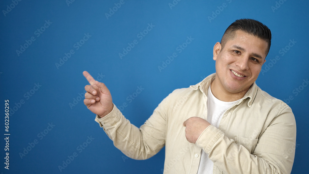 Young hispanic man smiling confident pointing to the side over isolated blue background Young hispanic man smiling confident pointing to the side over isolated blue background