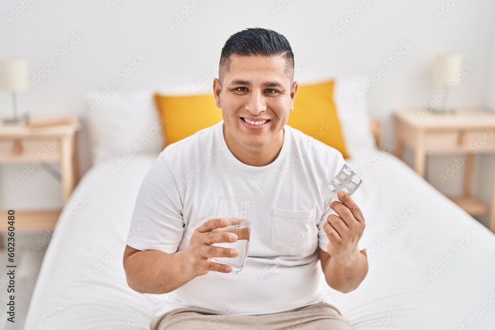Young latin man smiling confident holding pills and glass of water at bedroom