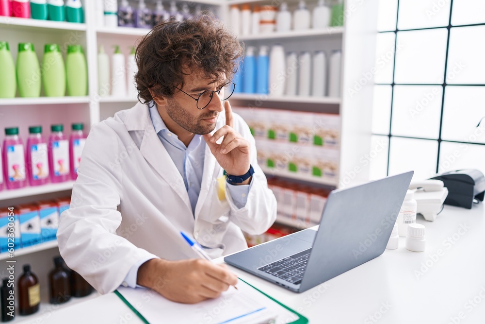 Young hispanic man pharmacist writing on document using laptop at pharmacy Young hispanic man pharmacist writing on document using laptop at pharmacy