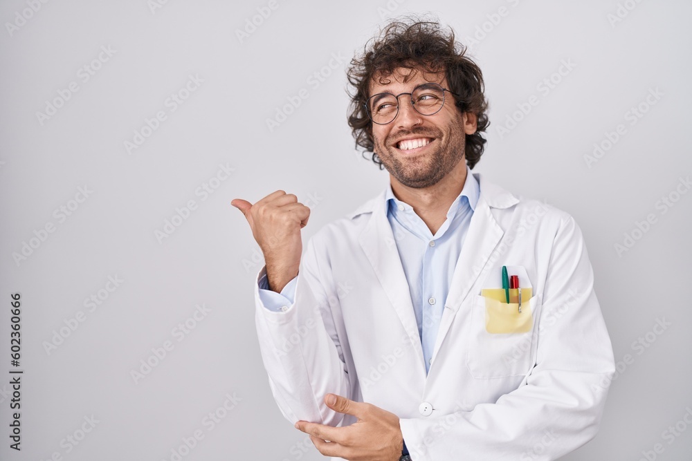 Hispanic young man wearing doctor uniform smiling with happy face looking and pointing to the side with thumb up. Hispanic young man wearing doctor uniform smiling with happy face looking and pointing to the side with thumb up.