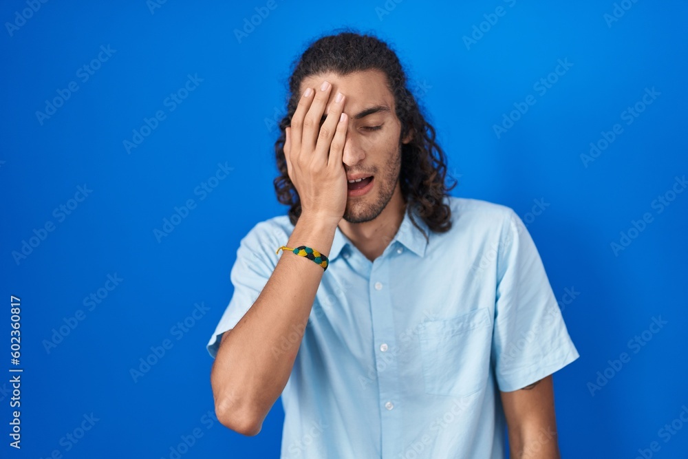 Young hispanic man standing over blue background yawning tired covering half face, eye and mouth with hand. face hurts in pain. Young hispanic man standing over blue background yawning tired covering half face, eye and mouth with hand. face hurts in pain.
