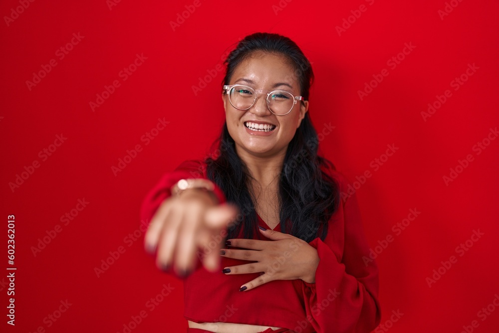 Asian young woman standing over red background laughing at you, pointing finger to the camera with hand over body, shame expression Asian young woman standing over red background laughing at you, pointing finger to the camera with hand over body, shame expression