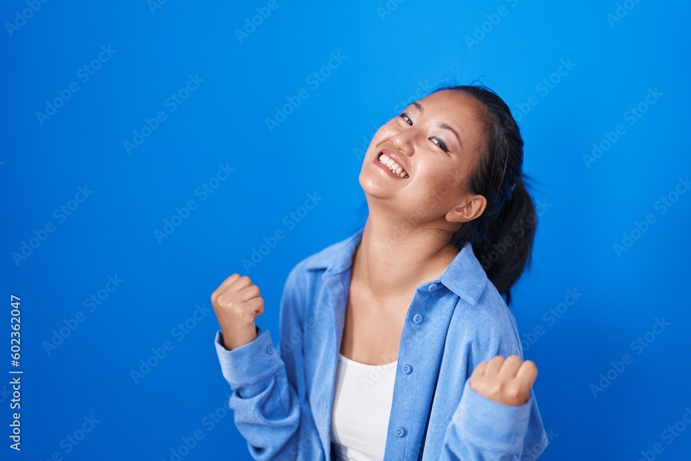 Asian young woman standing over blue background very happy and excited doing winner gesture with arms raised, smiling and screaming for success. celebration concept. Asian young woman standing over blue background very happy and excited doing winner gesture with arms raised, smiling and screaming for success. celebration concept.