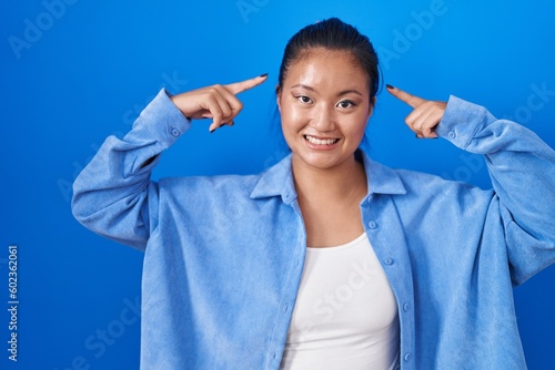Asian young woman standing over blue background smiling pointing to head with both hands finger, great idea or thought, good memory