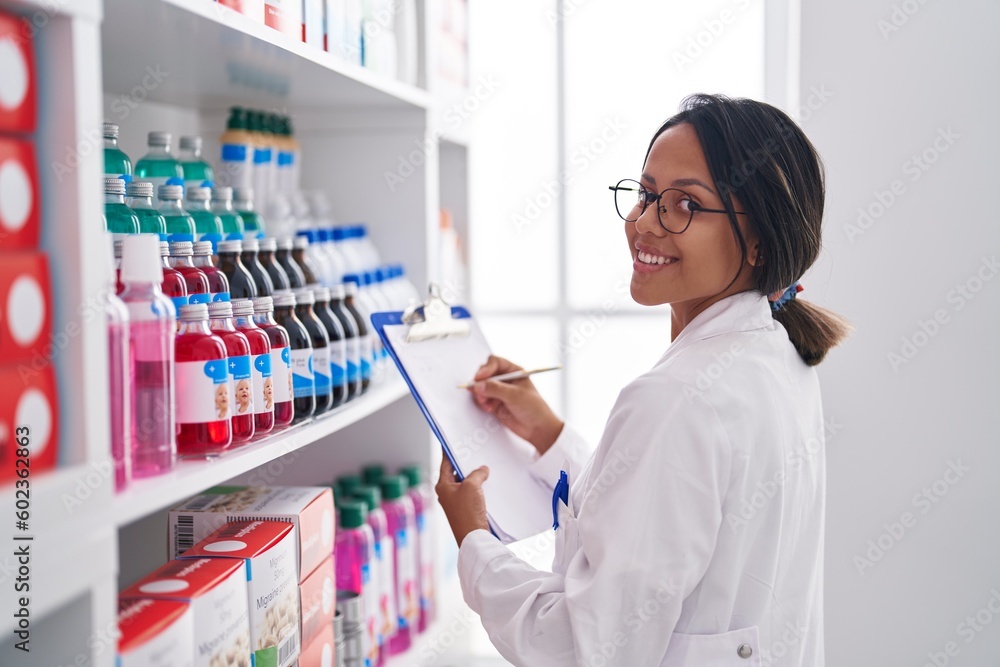 Young hispanic woman pharmacist writing on document looking shelving at pharmacy