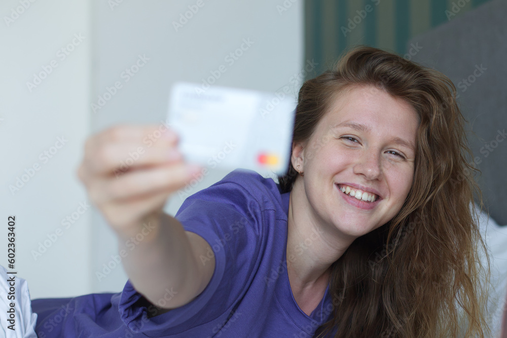 Young happy woman doing online shopping with smartphone, lay in bed in bedroom at home, hold in hand credit bank card and cell mobile phone and smile Young happy woman doing online shopping with smartphone, lay in bed in bedroom at home, hold in hand credit bank card and cell mobile phone and smile