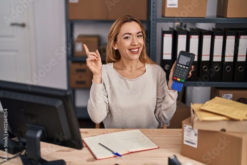 Hispanic woman working at small business ecommerce holding credit card and dataphone smiling happy pointing with hand and finger to the side