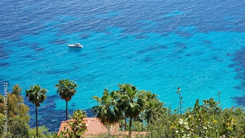 Small yacht on clear waters with green garden on the shore. French Riviera