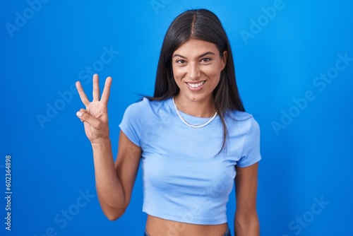 Brunette young woman standing over blue background showing and pointing up with fingers number three while smiling confident and happy.