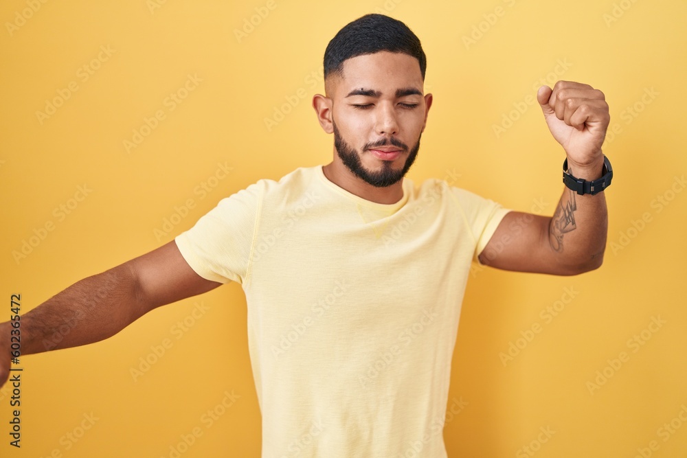 Young hispanic man standing over yellow background dancing happy and cheerful, smiling moving casual and confident listening to music Young hispanic man standing over yellow background dancing happy and cheerful, smiling moving casual and confident listening to music
