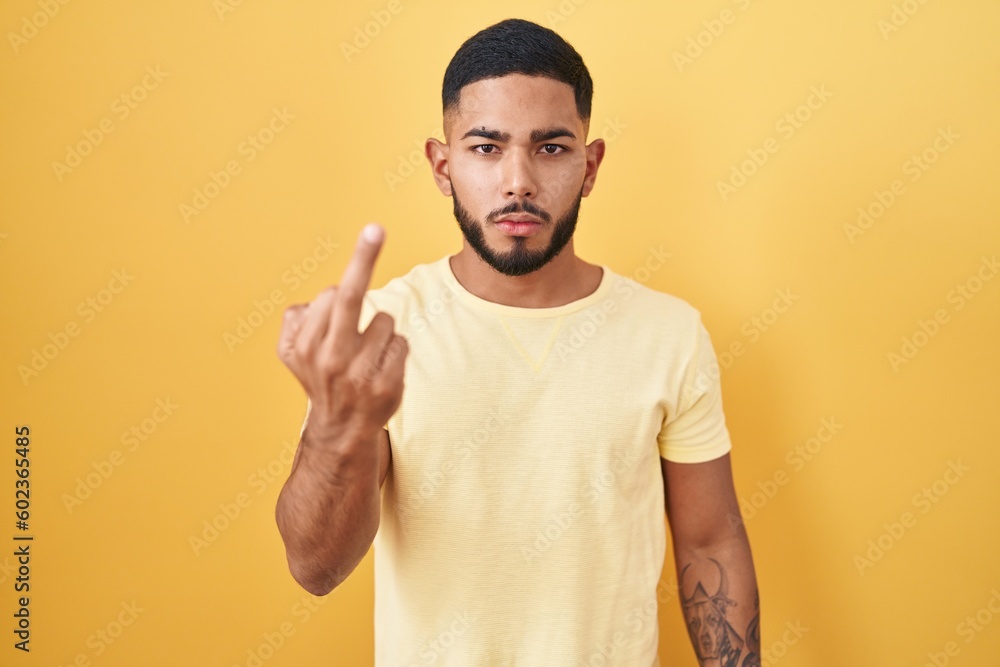 Young hispanic man standing over yellow background showing middle finger, impolite and rude fuck off expression Young hispanic man standing over yellow background showing middle finger, impolite and rude fuck off expression