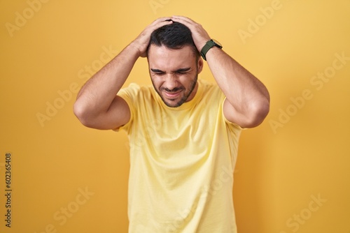 Young hispanic man standing over yellow background suffering from headache desperate and stressed because pain and migraine. hands on head.