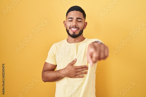 Young hispanic man standing over yellow background laughing at you, pointing finger to the camera with hand over body, shame expression
