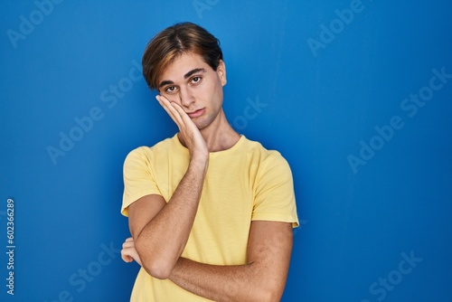 Young man standing over blue background thinking looking tired and bored with depression problems with crossed arms.
