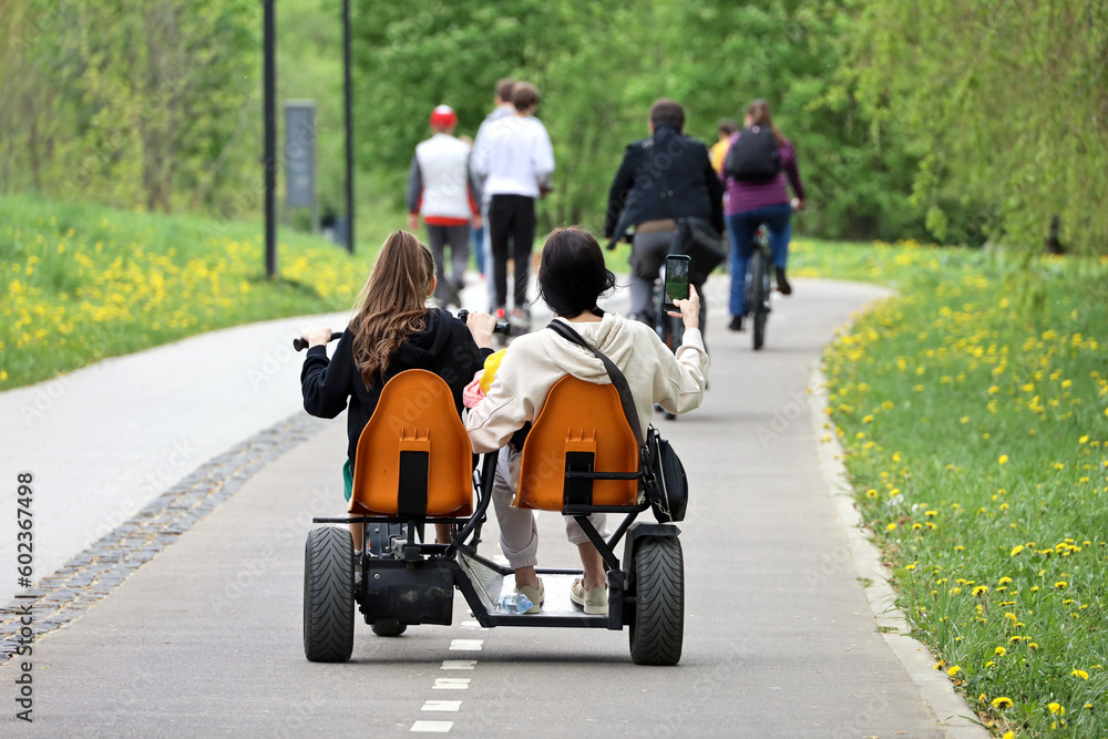 Two girls riding velomobile in a green park. Tandem bike renting for spring or summer leisure Two girls riding velomobile in a green park. Tandem bike renting for spring or summer leisure