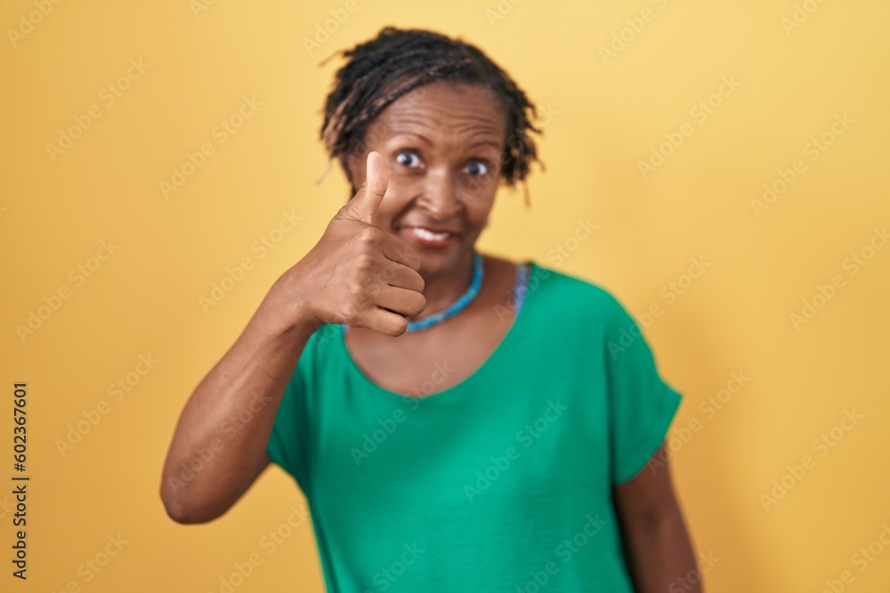 African woman with dreadlocks standing over yellow background doing happy thumbs up gesture with hand. approving expression looking at the camera showing success. African woman with dreadlocks standing over yellow background doing happy thumbs up gesture with hand. approving expression looking at the camera showing success.