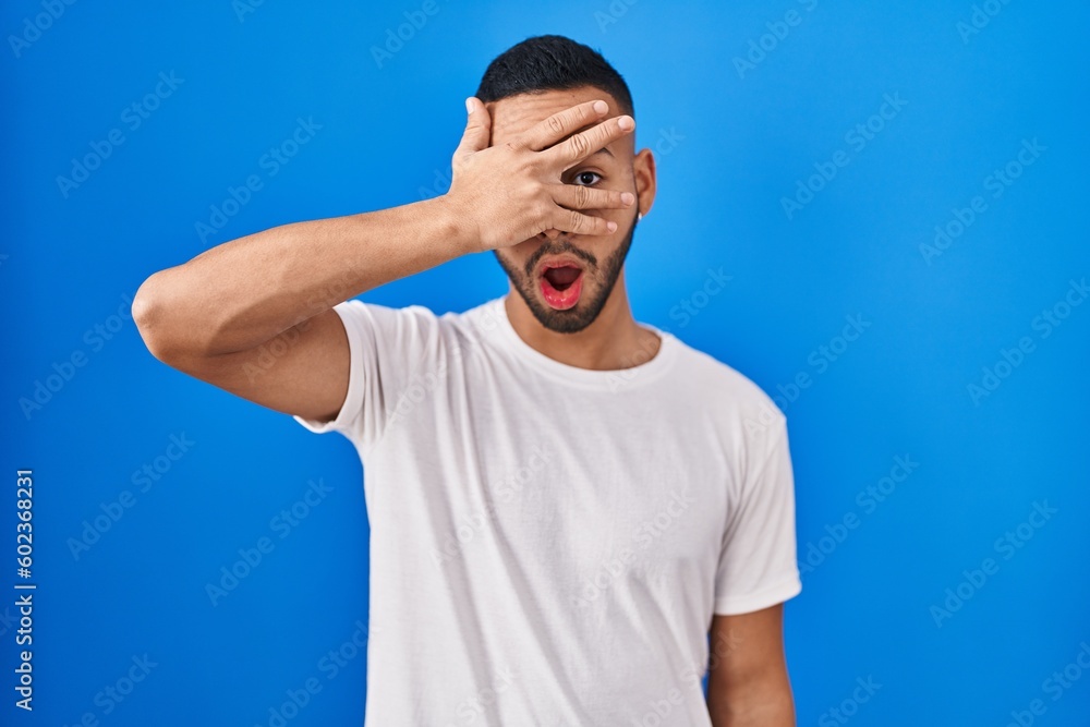 Young hispanic man standing over blue background peeking in shock covering face and eyes with hand, looking through fingers with embarrassed expression. Young hispanic man standing over blue background peeking in shock covering face and eyes with hand, looking through fingers with embarrassed expression.