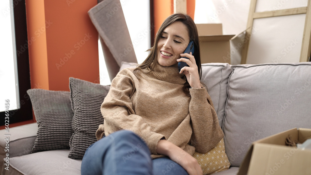 Beautiful hispanic woman talking on smartphone sitting on sofa at new home
