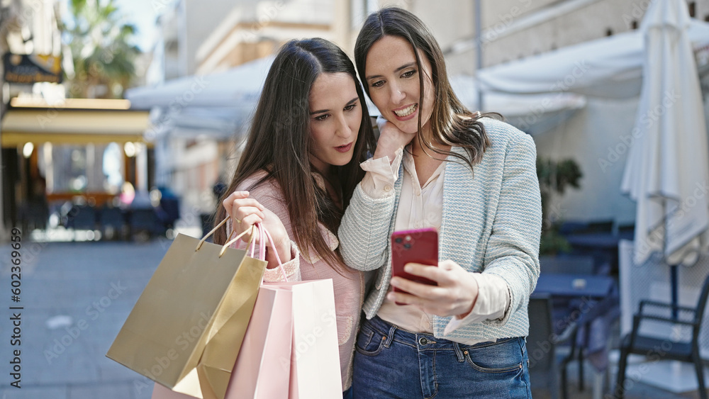 Two women going shopping holding bags using smartphone at coffee shop terrace Two women going shopping holding bags using smartphone at coffee shop terrace