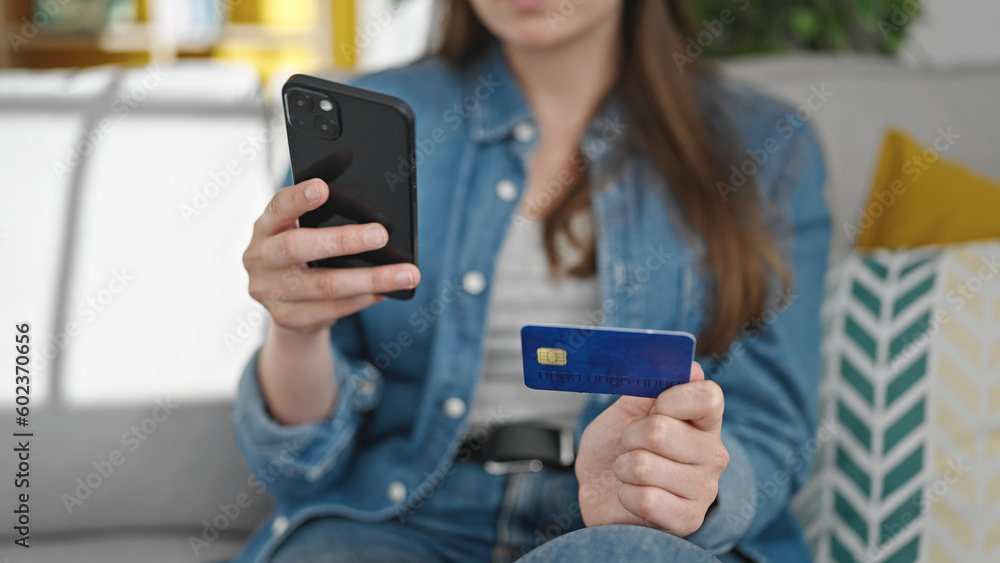 Young beautiful hispanic woman shopping with smartphone and credit card sitting on sofa at home