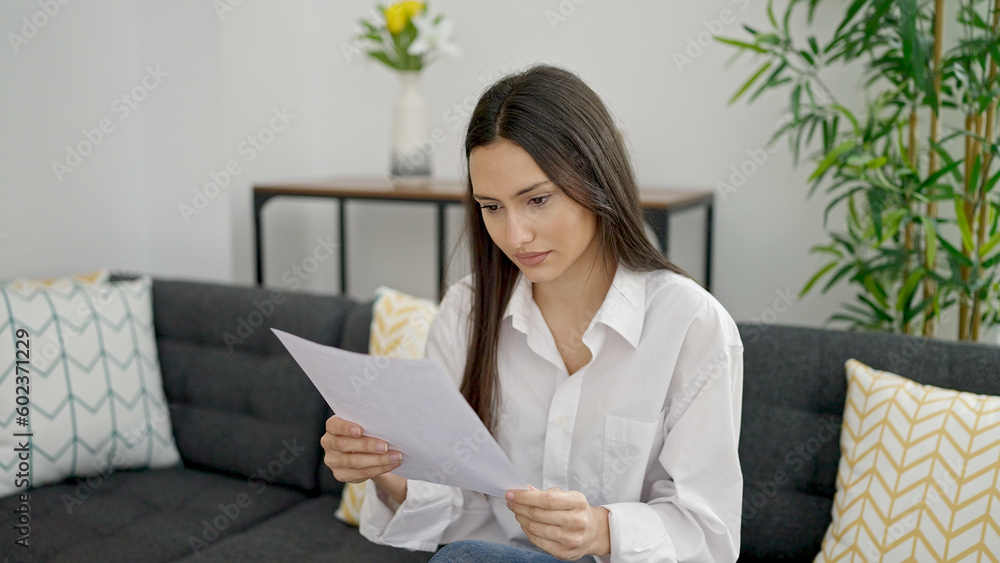 Fototapeta premium Young beautiful hispanic woman reading document sitting on sofa at home