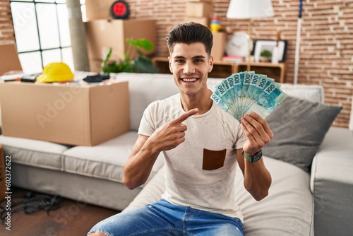 Young hispanic man moving to a new home holding brazilian reals smiling happy pointing with hand and finger