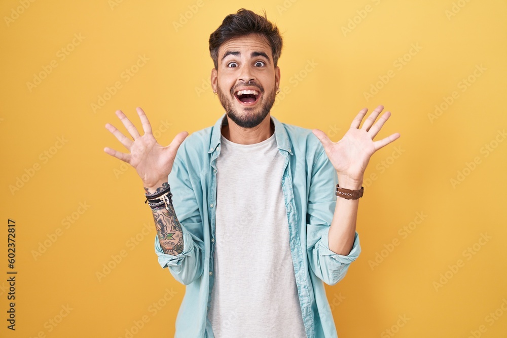 Young hispanic man with tattoos standing over yellow background celebrating crazy and amazed for success with arms raised and open eyes screaming excited. winner concept Young hispanic man with tattoos standing over yellow background celebrating crazy and amazed for success with arms raised and open eyes screaming excited. winner concept