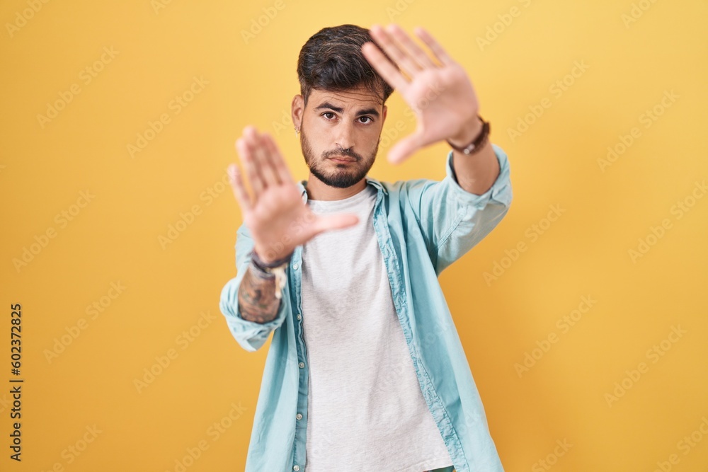 Young hispanic man with tattoos standing over yellow background doing frame using hands palms and fingers, camera perspective Young hispanic man with tattoos standing over yellow background doing frame using hands palms and fingers, camera perspective