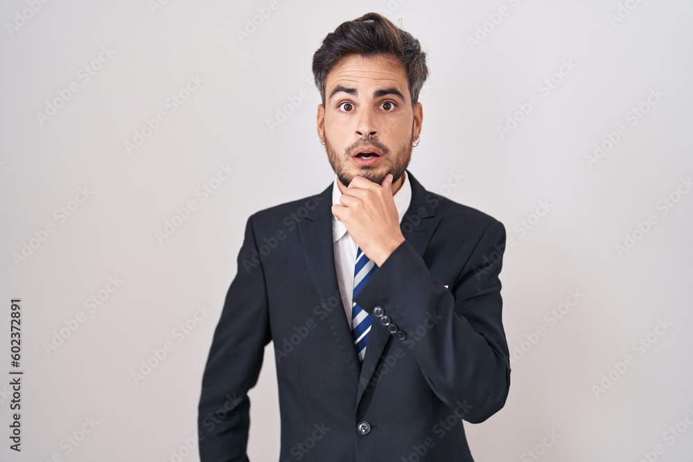 Young hispanic man with tattoos wearing business suit and tie looking fascinated with disbelief, surprise and amazed expression with hands on chin Young hispanic man with tattoos wearing business suit and tie looking fascinated with disbelief, surprise and amazed expression with hands on chin