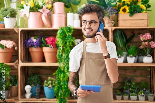 Young hispanic man florist talking on smartphone using touchpad at flower shop