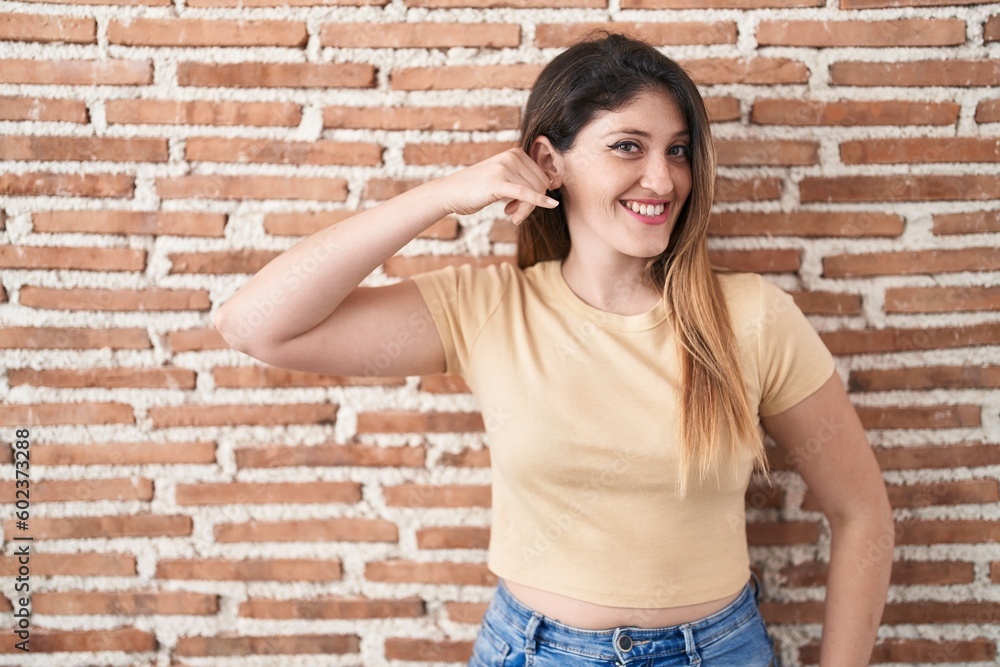 Young brunette woman standing over bricks wall smiling doing phone gesture with hand and fingers like talking on the telephone. communicating concepts. Young brunette woman standing over bricks wall smiling doing phone gesture with hand and fingers like talking on the telephone. communicating concepts.