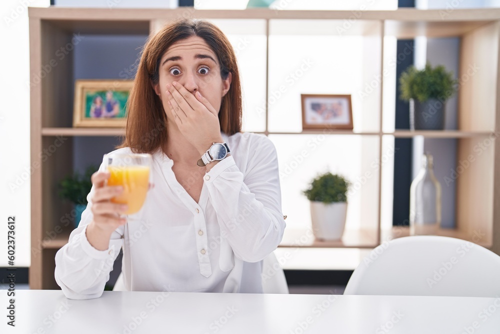 Brunette woman drinking glass of orange juice shocked covering mouth with hands for mistake. secret concept. Brunette woman drinking glass of orange juice shocked covering mouth with hands for mistake. secret concept.