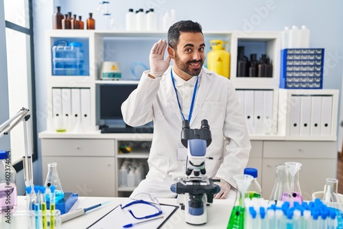 Young hispanic man with beard working at scientist laboratory smiling with hand over ear listening an hearing to rumor or gossip. deafness concept.