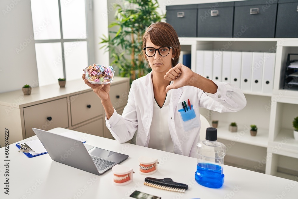 Brunette dentist woman holding sweets with angry face, negative sign showing dislike with thumbs down, rejection concept Brunette dentist woman holding sweets with angry face, negative sign showing dislike with thumbs down, rejection concept