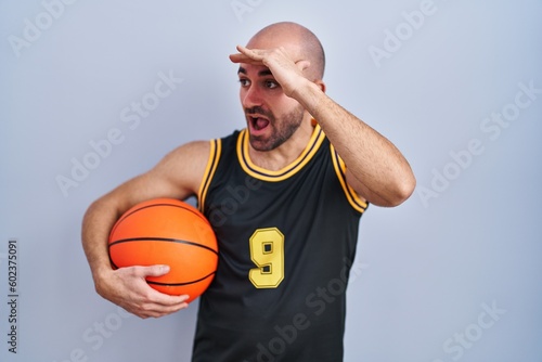 Young bald man with beard wearing basketball uniform holding ball very happy and smiling looking far away with hand over head. searching concept.