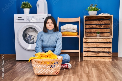 Young hispanic woman at laundry room thinking attitude and sober expression looking self confident