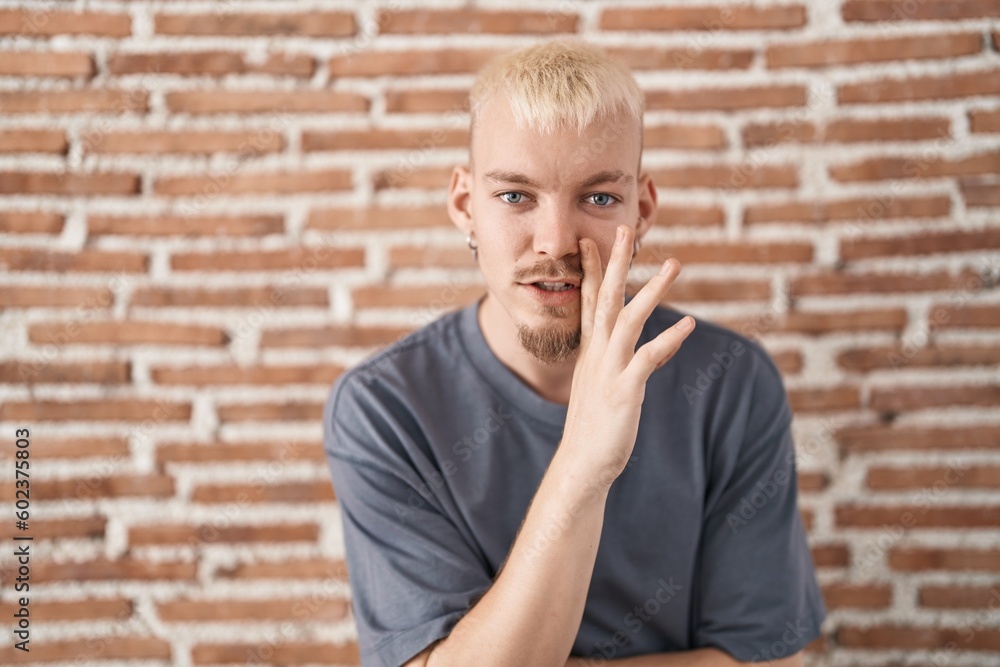 Young caucasian man standing over bricks wall hand on mouth telling secret rumor, whispering malicious talk conversation Young caucasian man standing over bricks wall hand on mouth telling secret rumor, whispering malicious talk conversation