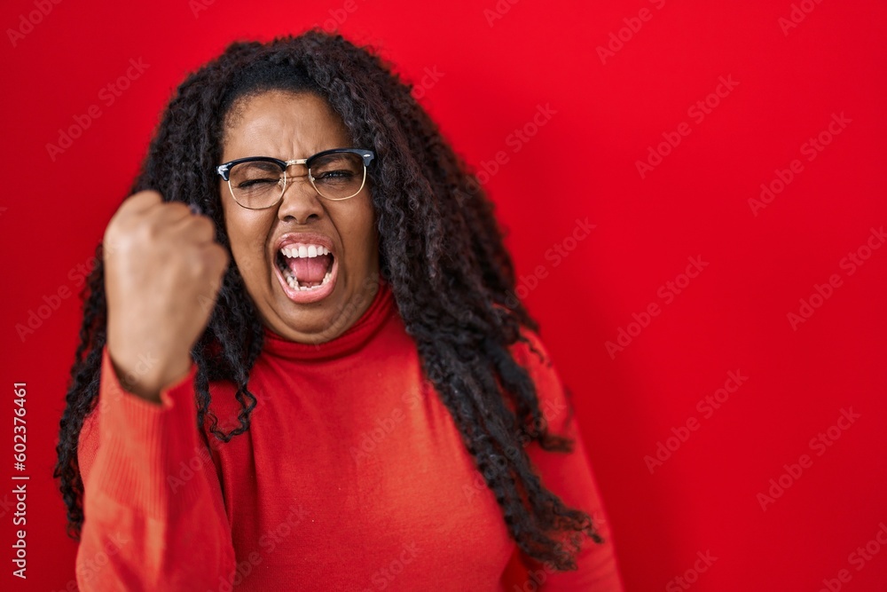 Plus size hispanic woman standing over red background angry and mad raising fist frustrated and furious while shouting with anger. rage and aggressive concept. Plus size hispanic woman standing over red background angry and mad raising fist frustrated and furious while shouting with anger. rage and aggressive concept.