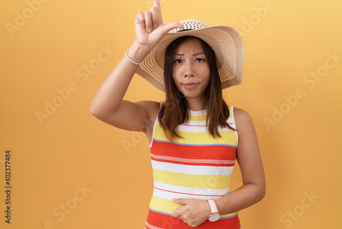 Middle age chinese woman wearing summer hat over yellow background making fun of people with fingers on forehead doing loser gesture mocking and insulting.