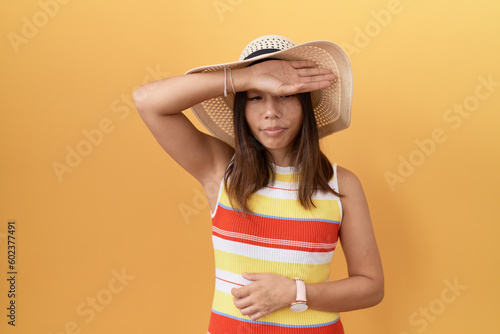 Middle age chinese woman wearing summer hat over yellow background covering eyes with arm, looking serious and sad. sightless, hiding and rejection concept