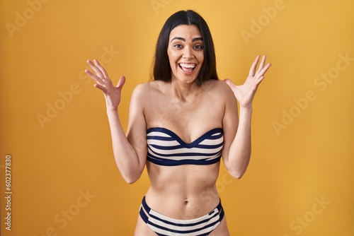 Young brunette woman wearing bikini over yellow background celebrating mad and crazy for success with arms raised and closed eyes screaming excited. winner concept