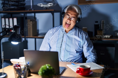 Hispanic senior man wearing call center agent headset at night sticking tongue out happy with funny expression. emotion concept.