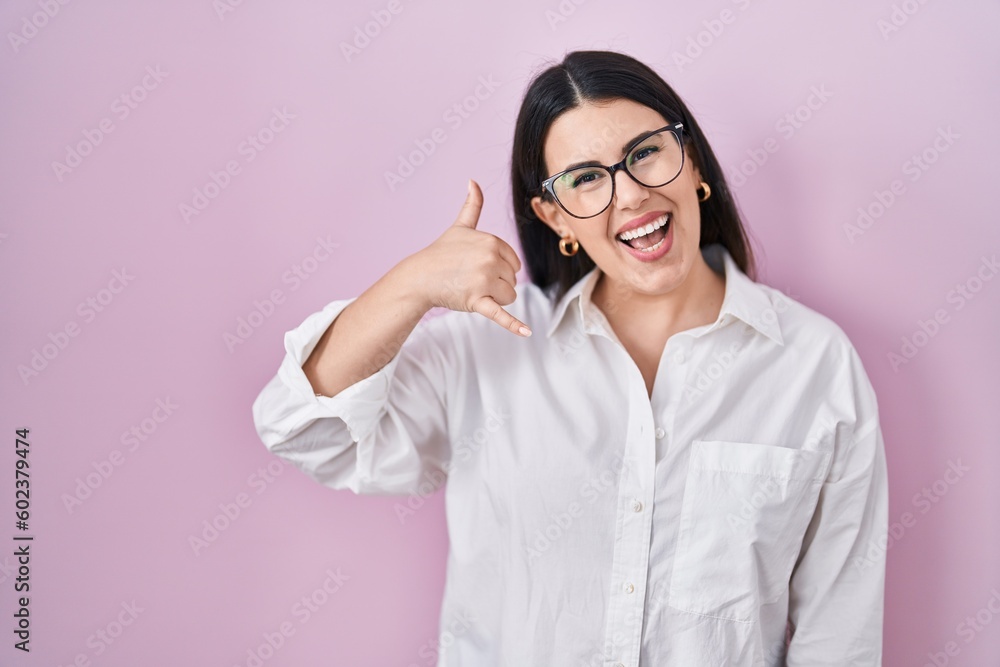 Young brunette woman standing over pink background smiling doing phone gesture with hand and fingers like talking on the telephone. communicating concepts.