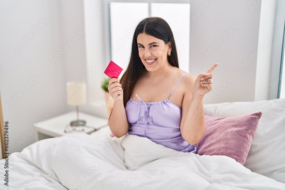Young brunette woman wearing lingerie and holding condom on the bed smiling happy pointing with hand and finger to the side