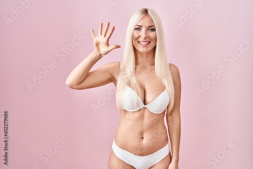 Caucasian woman wearing lingerie over pink background showing and pointing up with fingers number five while smiling confident and happy.
