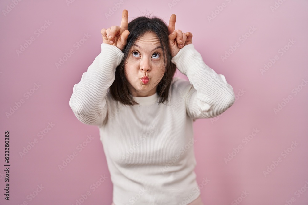 Woman with down syndrome standing over pink background doing funny gesture with finger over head as bull horns Woman with down syndrome standing over pink background doing funny gesture with finger over head as bull horns