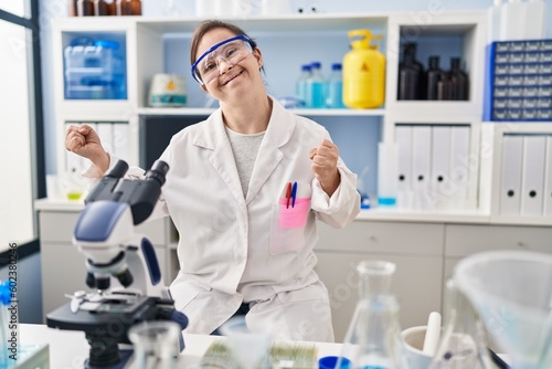 Hispanic girl with down syndrome working at scientist laboratory very happy and excited doing winner gesture with arms raised, smiling and screaming for success. celebration concept.